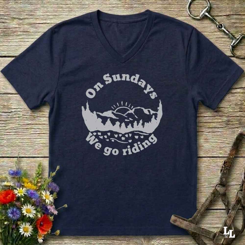 A navy blue V-neck t-shirt with a white graphic reading 'On Sundays We go riding' around a mountain and tree silhouette, laid flat on a rustic wood background with flowers and horse tack props.