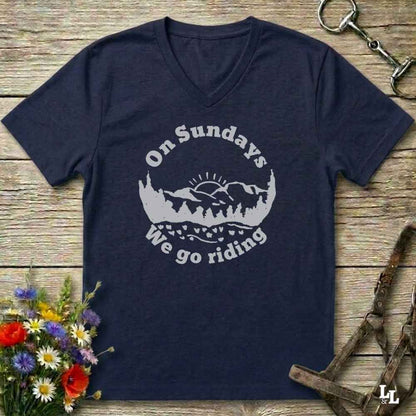 A navy blue V-neck t-shirt with a white graphic reading 'On Sundays We go riding' around a mountain and tree silhouette, laid flat on a rustic wood background with flowers and horse tack props.