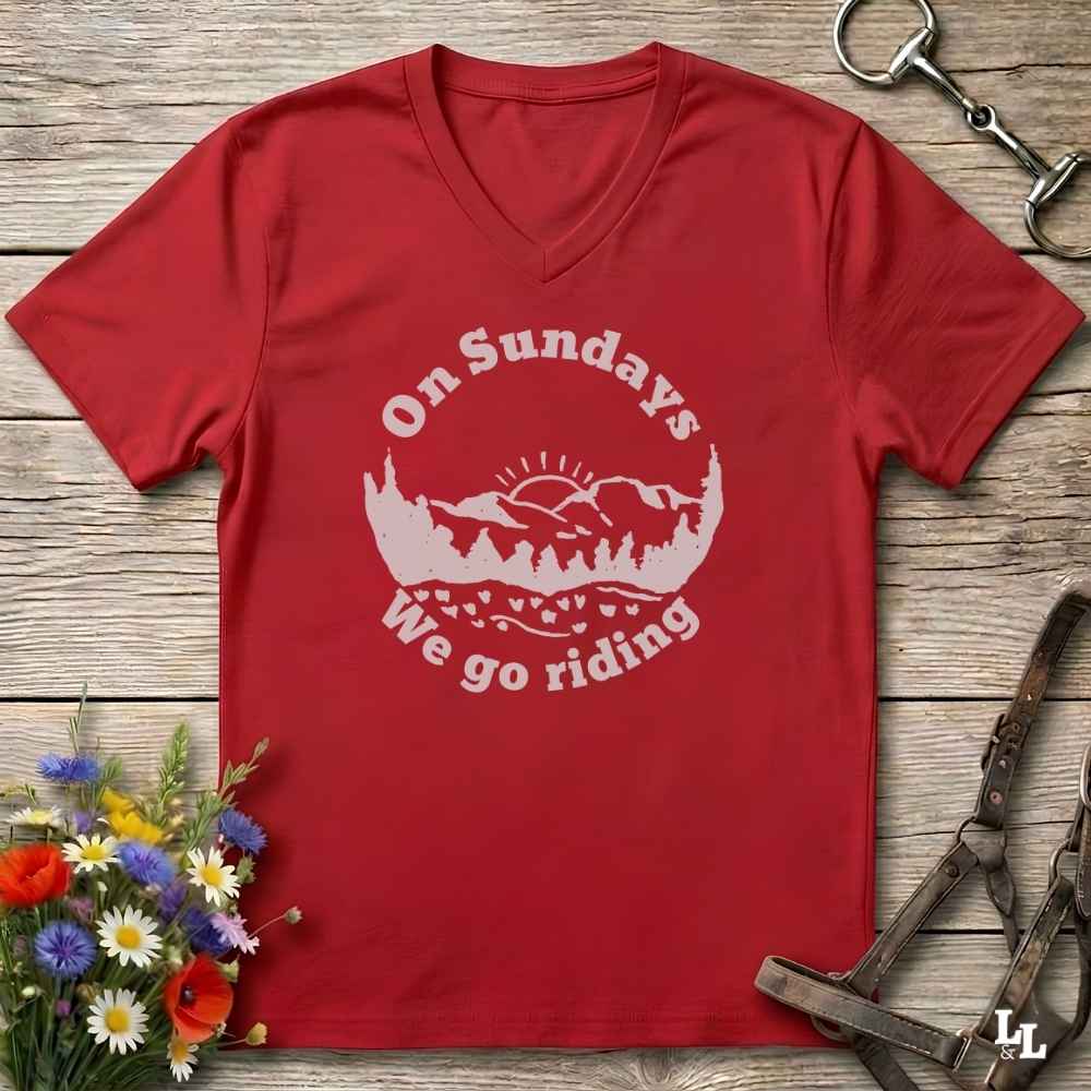 A red V-neck t-shirt with a white graphic reading 'On Sundays We go riding' over a mountain and tree silhouette, laid flat on a rustic wood surface with flowers and horse tack props.