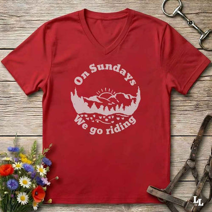 A red V-neck t-shirt with a white graphic reading 'On Sundays We go riding' over a mountain and tree silhouette, laid flat on a rustic wood surface with flowers and horse tack props.