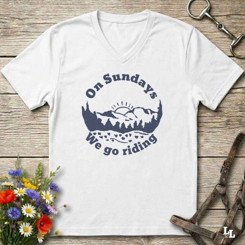A white v-neck t-shirt with a navy blue graphic reading 'On Sundays We go riding' around a mountain and tree silhouette, laid flat on a rustic wooden surface with flowers and horse tack props.