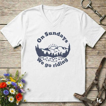 A white v-neck t-shirt with a navy blue graphic reading 'On Sundays We go riding' around a mountain and tree silhouette, laid flat on a rustic wooden surface with flowers and horse tack props.