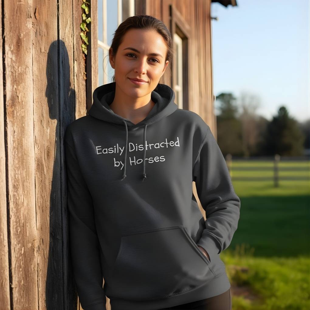 Person wearing a hoodie with 'Easily Distracted by Horses' text, standing in front of a wooden building. Easily Distracted by Horses Hoodie for Horse Riders, Barn Wear Riding Apparel