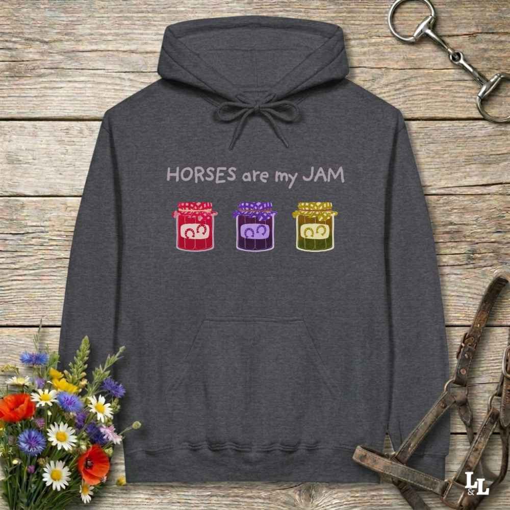 A dark gray pullover hoodie laid flat on a rustic wooden surface, featuring the text 'HORSES are my JAM' above three colorful jars with horse faces inside. The hoodie is styled with a bouquet of flowers, horse tack, and a leather strap around it.