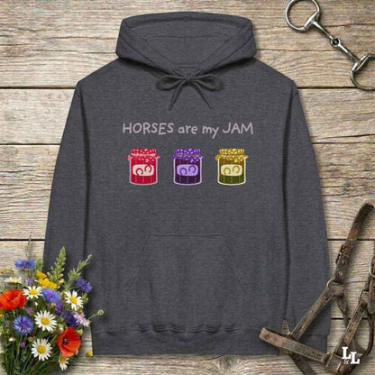 A dark gray pullover hoodie laid flat on a rustic wooden surface, featuring the text 'HORSES are my JAM' above three colorful jars with horse faces inside. The hoodie is styled with a bouquet of flowers, horse tack, and a leather strap around it.