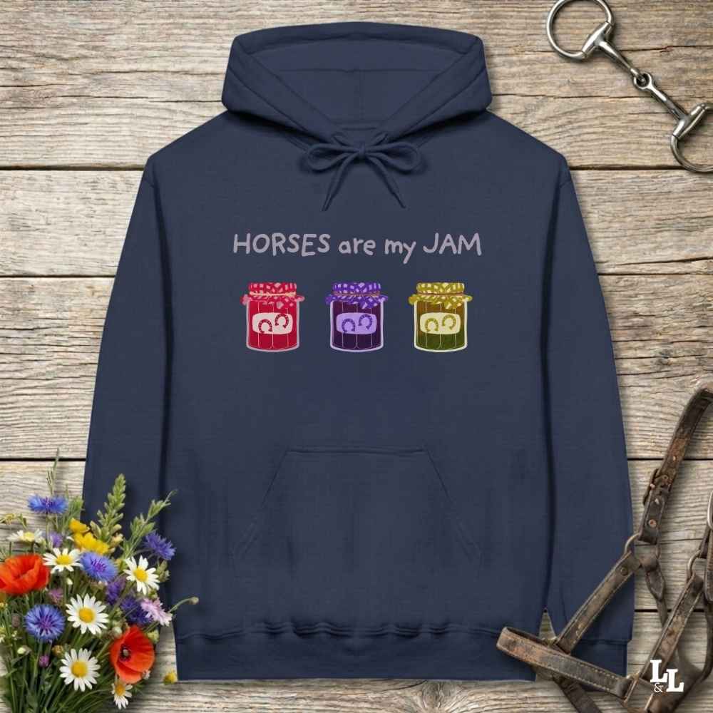 A navy blue hoodie laid flat on a rustic wood background, with the text 'HORSES are my JAM' and three colorful horse-themed jar graphics on the chest. The hoodie is styled with equestrian accessories and flowers around it.