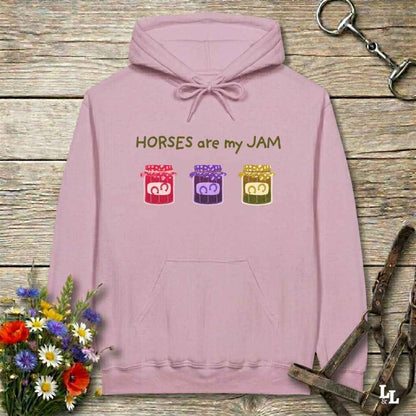 A light pink pullover hoodie laid flat on a rustic wooden surface, featuring the text 'HORSES are my JAM' above three colorful cartoon horse heads in red, purple, and green. The hoodie is styled with a bouquet of flowers, horse tack, and a leather strap around it.