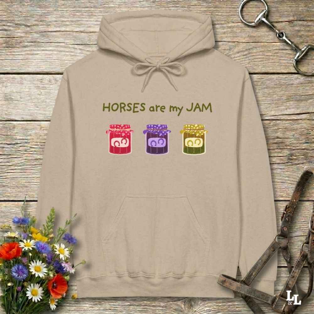 A beige pullover hoodie with the text 'HORSES are my JAM' and three colorful horse head graphics, laid flat on a rustic wood background with flowers and horse tack props.