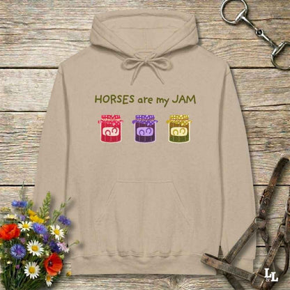 A beige pullover hoodie with the text 'HORSES are my JAM' and three colorful horse head graphics, laid flat on a rustic wood background with flowers and horse tack props.