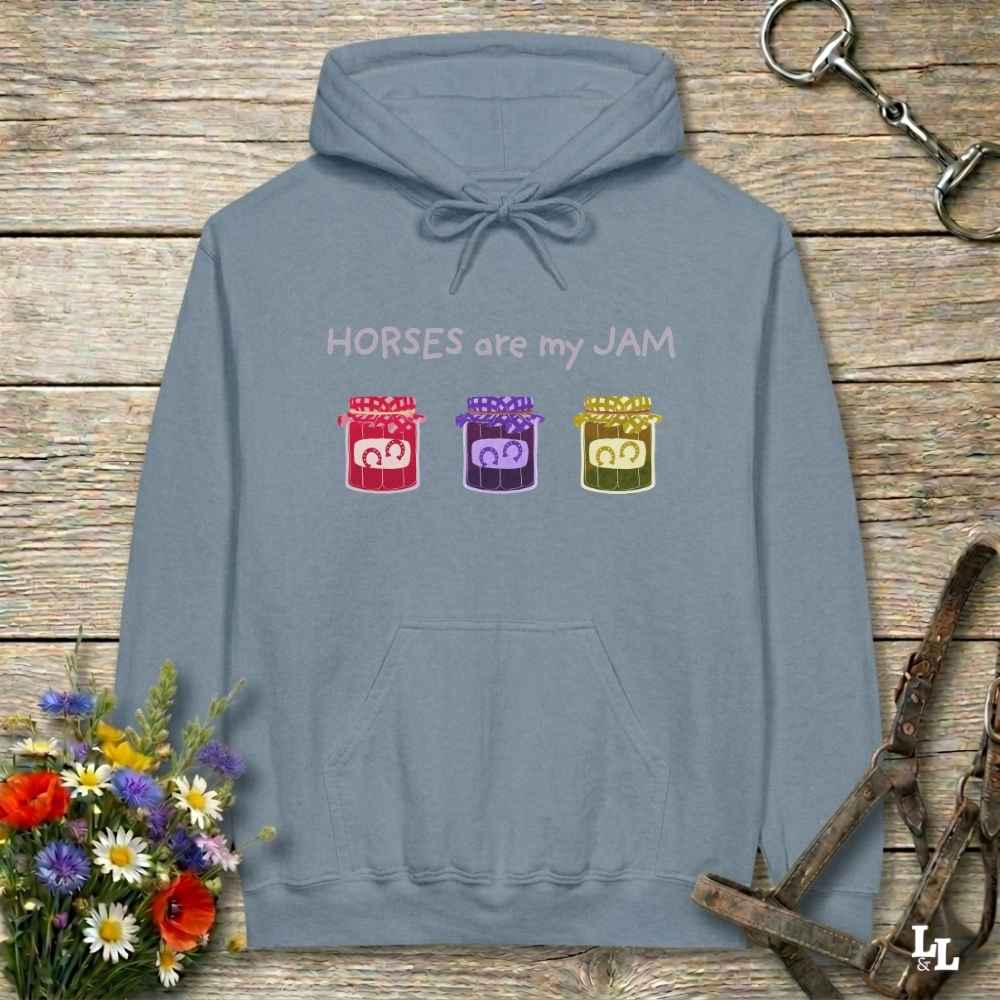 A slate blue hoodie with a graphic print that reads 'HORSES are my JAM' above three colorful jars of horse treats, displayed on a rustic wooden background with decorative flowers and a horse bridle.