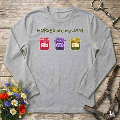 A light gray long-sleeve t-shirt with a graphic print reading 'HORSES are my JAM' above three colorful jars of horse manure. The shirt is displayed in a flat-lay arrangement on a rustic wooden surface, surrounded by equestrian-themed props including a leather bridle, a horse bit, and a small bouquet of wildflowers.