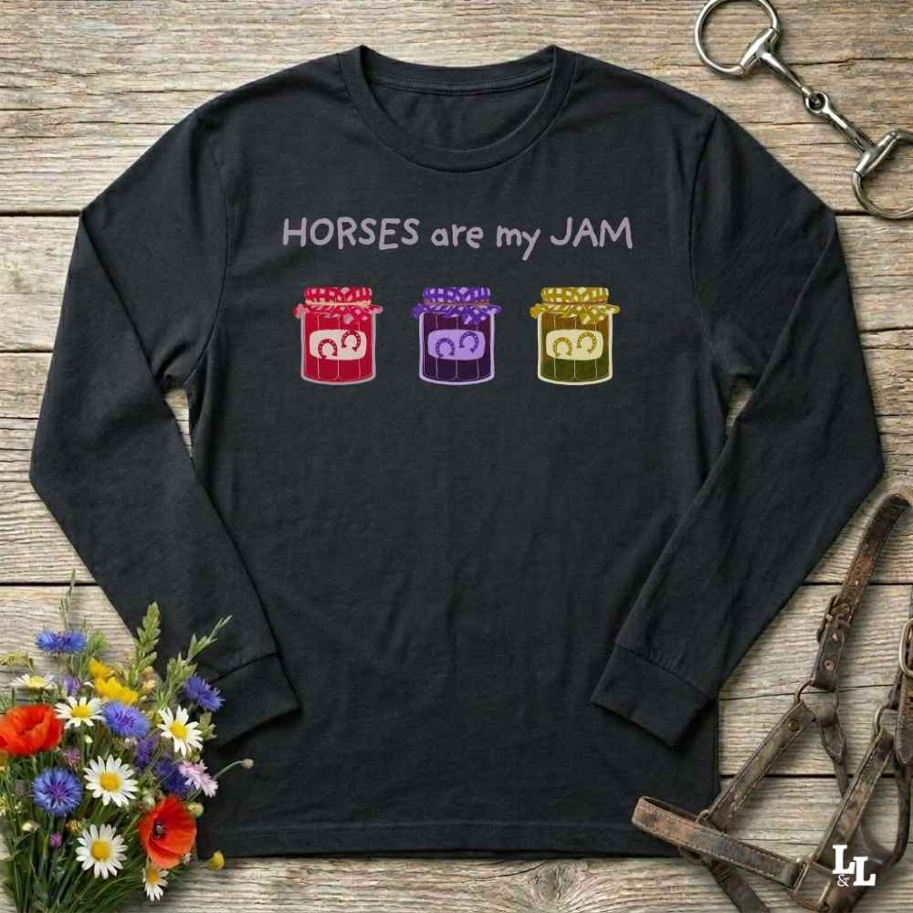 A black long-sleeve t-shirt with the text 'HORSES are my JAM' and three colorful horse silhouettes in red, purple, and gold, displayed on a rustic wooden background with equestrian accessories and flowers.