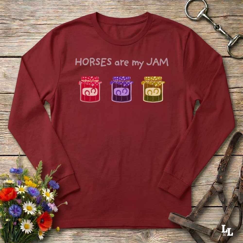 A long-sleeve crewneck t-shirt in maroon red, laid flat on a rustic wooden surface. The shirt features the text 'HORSES are my JAM' in white above three colorful jars containing cartoon horses (red, purple, and gold).