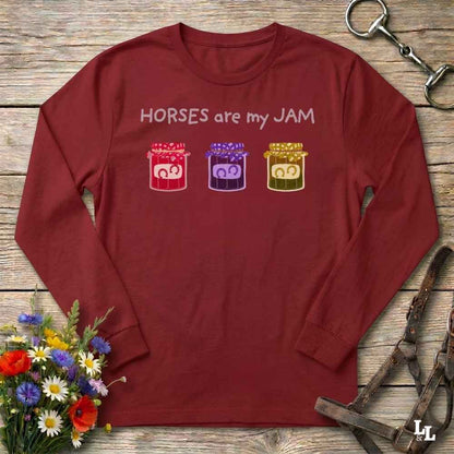 A long-sleeve crewneck t-shirt in maroon red, laid flat on a rustic wooden surface. The shirt features the text 'HORSES are my JAM' in white above three colorful jars containing cartoon horses (red, purple, and gold).