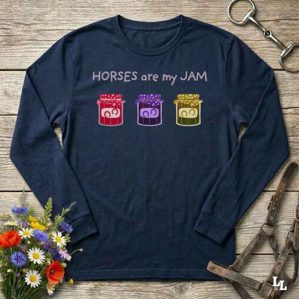 A navy blue long-sleeve t-shirt with a graphic print that reads 'HORSES are my JAM' above three colorful jars of horse manure. The shirt is displayed in a flat-lay arrangement on a rustic wooden background, styled with a bouquet of wildflowers, a horse bit, and a leather bridle.