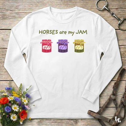 A white long-sleeve t-shirt with a graphic print reading 'HORSES are my JAM' above three colorful horse heads, laid flat on a rustic wooden surface with equestrian-themed props.