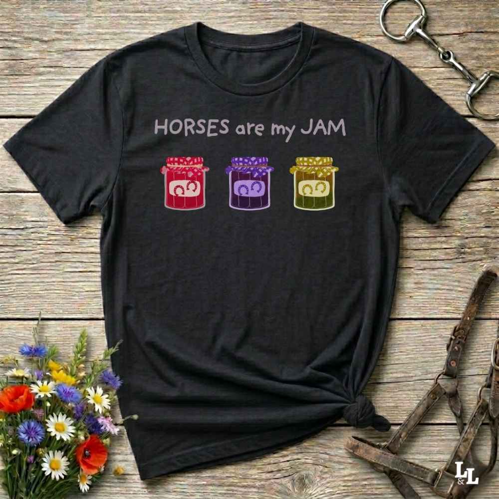 A black graphic t-shirt laid flat on a rustic wooden surface, styled with equestrian-themed props including a halter, horse boots, and a small bouquet of wildflowers. The shirt features the text 'HORSES are my JAM' above three colorful horse silhouettes in red, purple, and yellow.