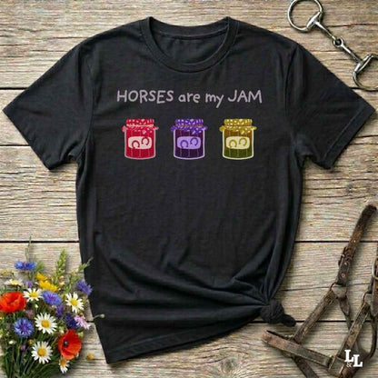 A black graphic t-shirt laid flat on a rustic wooden surface, styled with equestrian-themed props including a halter, horse boots, and a small bouquet of wildflowers. The shirt features the text 'HORSES are my JAM' above three colorful horse silhouettes in red, purple, and yellow.