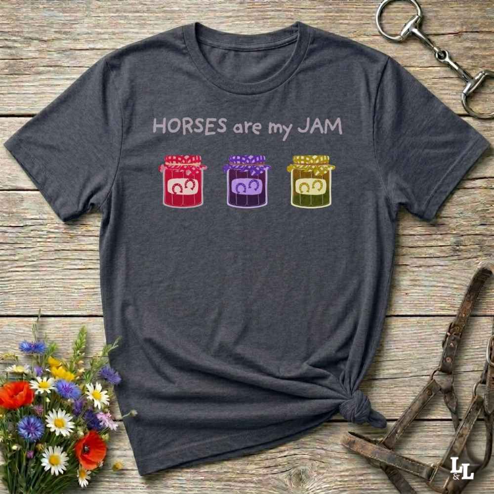A dark gray graphic t-shirt laid flat on a rustic wooden surface, styled with equestrian-themed props including a halter, horse boots, and a small bouquet of wildflowers. The shirt features the text 'HORSES are my JAM' above three colorful horse silhouettes inside red, purple, and yellow stables.
