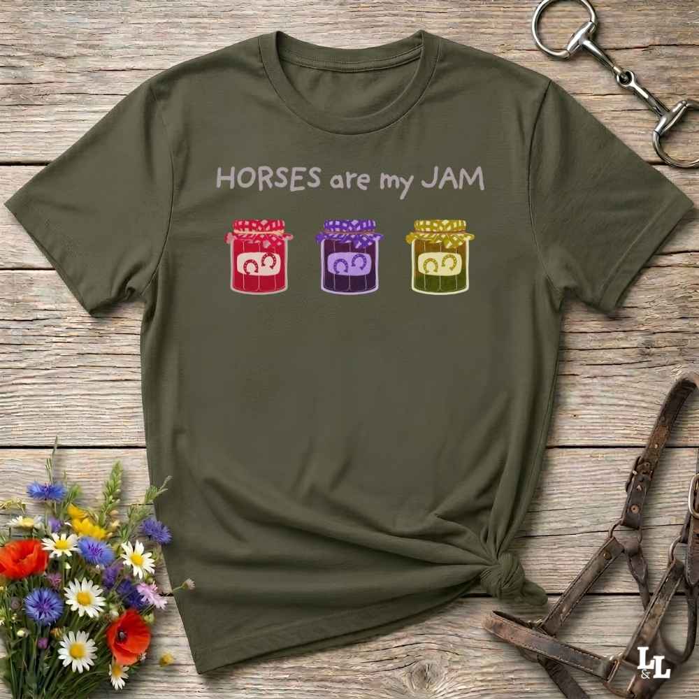 An olive green graphic t-shirt laid flat on a rustic wooden surface, styled with equestrian accessories and flowers. The shirt features the text 'HORSES are my JAM' above three colorful horse silhouettes in red, purple, and yellow.
