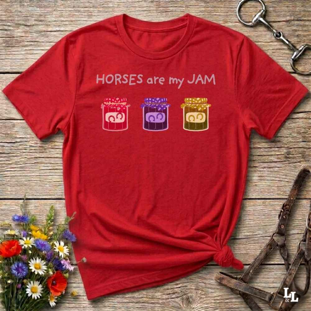 A red short-sleeve graphic T-shirt laid flat on a rustic wooden surface, with the text 'HORSES are my JAM' above three small horse silhouettes in pink, purple, and yellow. The shirt is styled with flowers, a halter, and a metal tack piece around it.