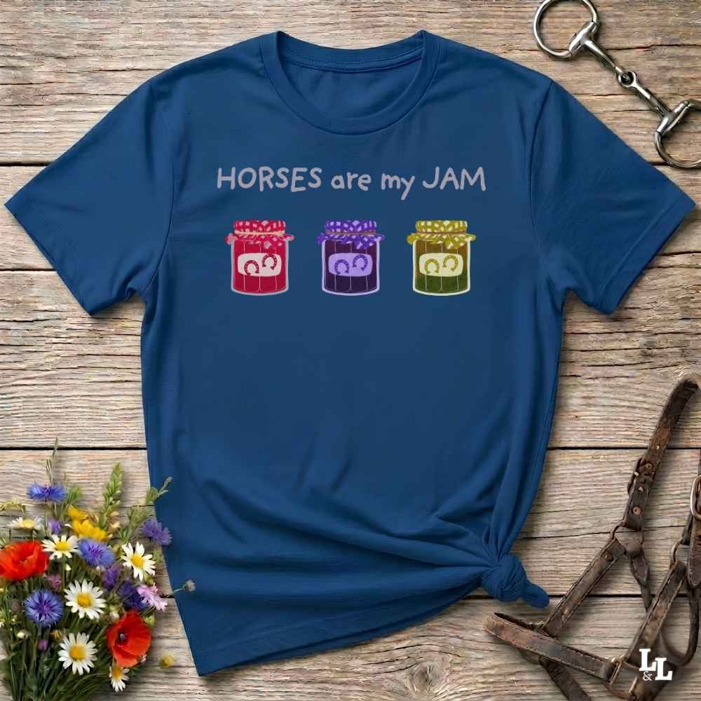 A navy blue graphic t-shirt laid flat on a rustic wooden surface, styled with a horse bit in the top right, a leather halter in the bottom right, and a small bouquet of colorful wildflowers in the bottom left. The t-shirt features the text 'HORSES are my JAM' above three small jars of horse manure in red, purple, and green.