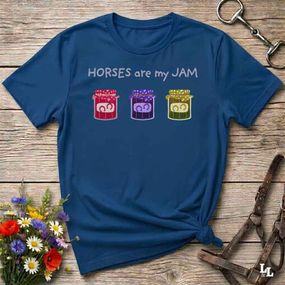 A navy blue graphic t-shirt laid flat on a rustic wooden surface, styled with a horse bit in the top right, a leather halter in the bottom right, and a small bouquet of colorful wildflowers in the bottom left. The t-shirt features the text 'HORSES are my JAM' above three small jars of horse manure in red, purple, and green.