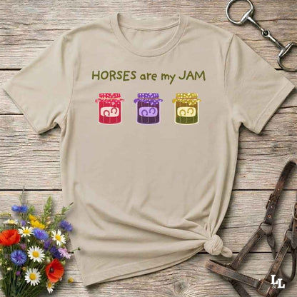 A beige graphic t-shirt laid flat on a rustic wooden surface, styled with equestrian accessories and a small bouquet of flowers. The shirt features the text 'HORSES are my JAM' in olive green above three colorful cartoon horse silhouettes inside red, purple, and yellow jars.
