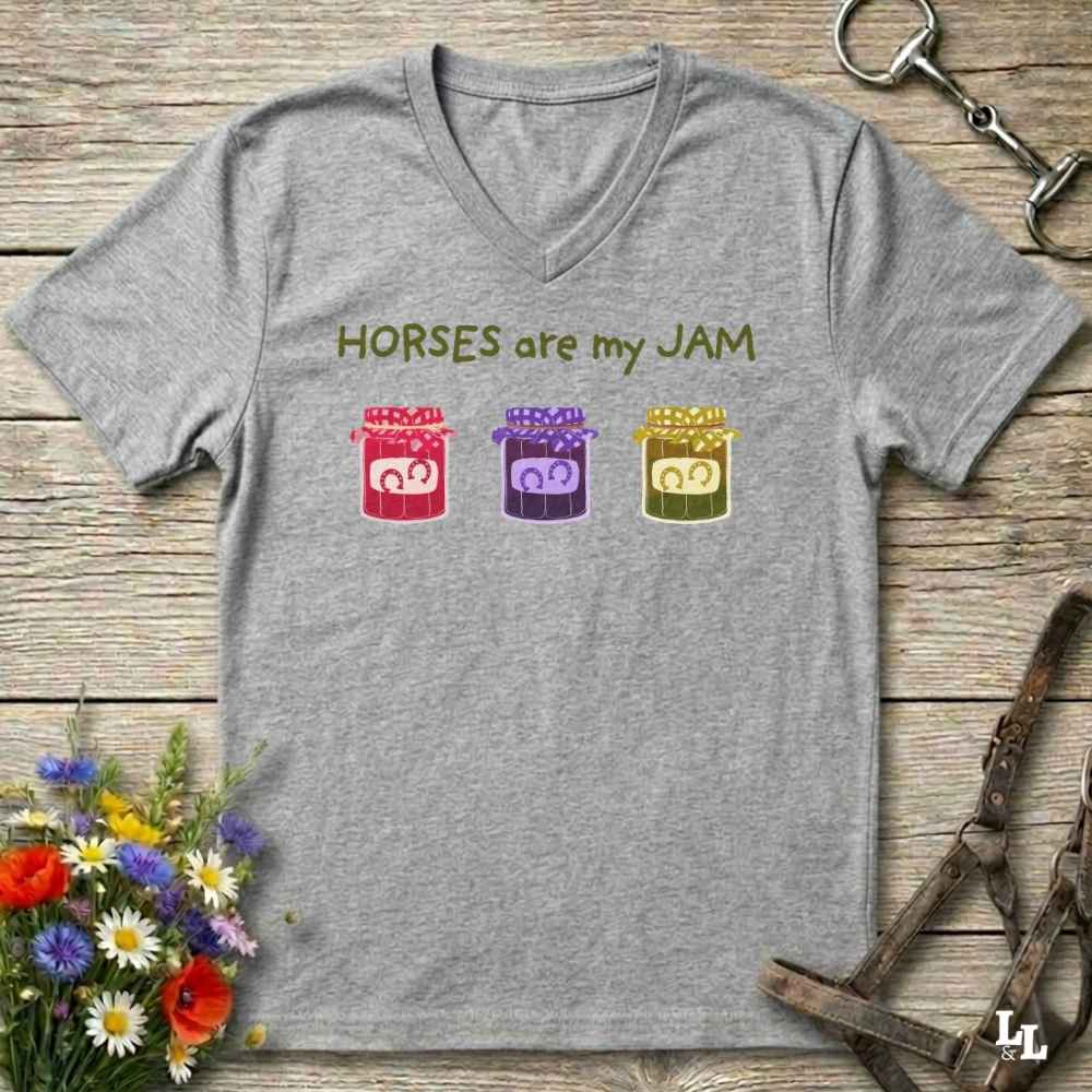 A heather gray V-neck t-shirt laid flat on a rustic wooden surface, featuring the text 'HORSES are my JAM' above three colorful cartoon jars with horse faces. The shirt is styled with equestrian and floral props.