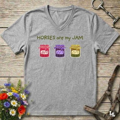 A heather gray V-neck t-shirt laid flat on a rustic wooden surface, featuring the text 'HORSES are my JAM' above three colorful cartoon jars with horse faces. The shirt is styled with equestrian and floral props.