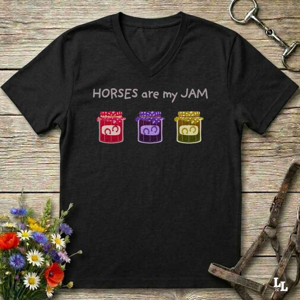 A black V-neck t-shirt laid flat on a rustic wooden surface, featuring the text 'HORSES are my JAM' above three colorful cartoon jars with horse faces. The shirt is styled with a bouquet of wildflowers, a horse bit, and a leather bridle.