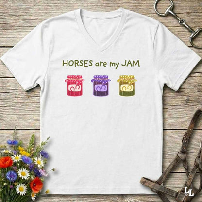 A white V-neck t-shirt laid flat on a rustic wooden surface, featuring the text 'HORSES are my JAM' above three colorful cartoon jars with horse faces. The shirt is styled with equestrian-themed props including a leather bridle, horse tack, and a small bouquet of wildflowers.