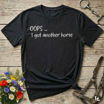 A black short-sleeve graphic T-shirt laid flat on a rustic wooden surface, with white text reading 'OOPS .... I got another horse'