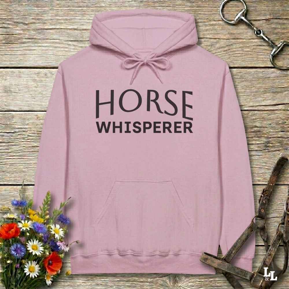 A dusty pink pullover hoodie with 'HORSE WHISPERER' printed in black block letters on the chest, displayed on a rustic wooden background with equestrian-themed props.