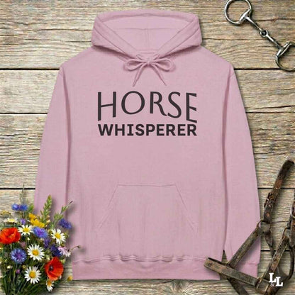 A dusty pink pullover hoodie with 'HORSE WHISPERER' printed in black block letters on the chest, displayed on a rustic wooden background with equestrian-themed props.