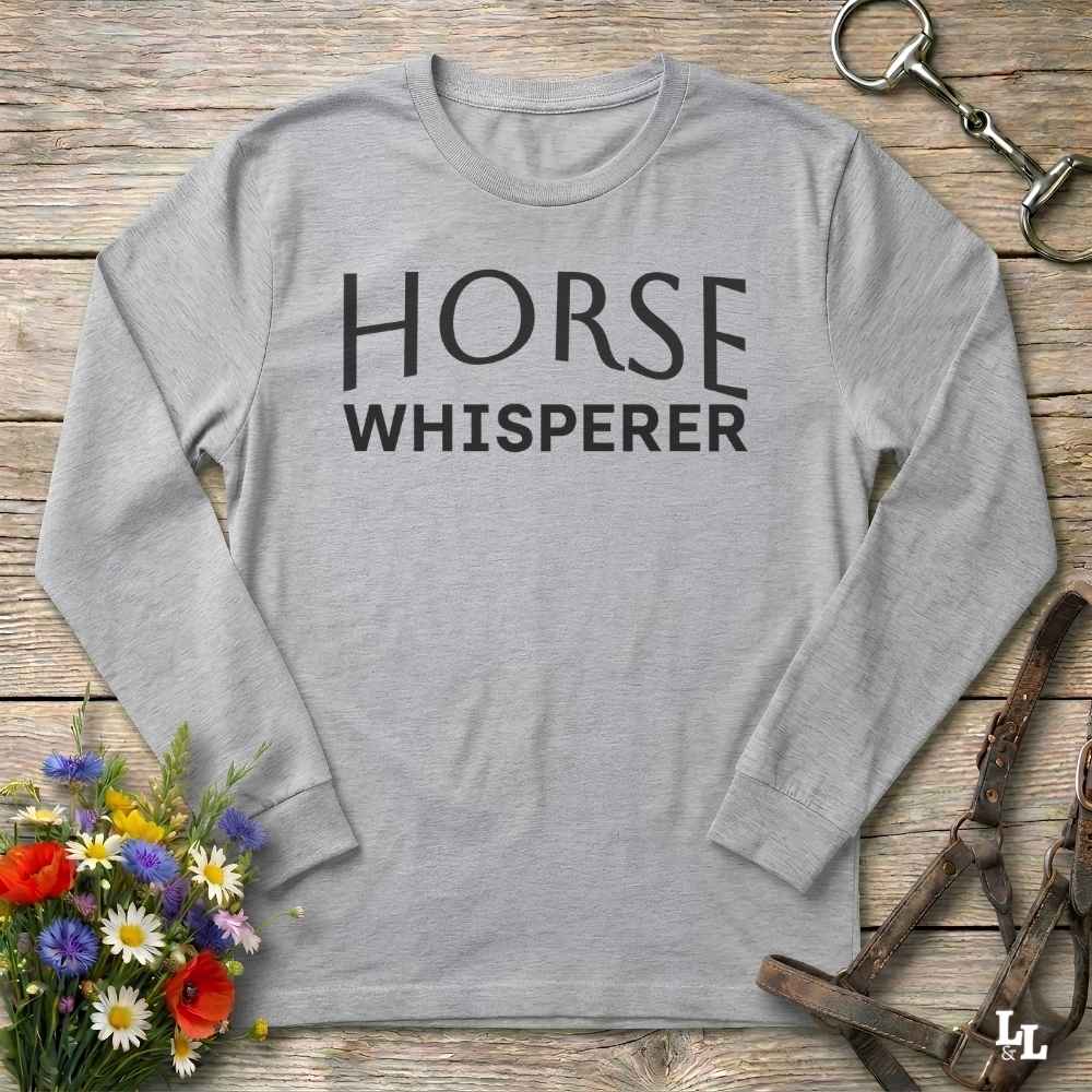 A light gray long-sleeve t-shirt with 'HORSE WHISPERER' printed in black text on the chest, styled in a flat-lay on a rustic wooden background with equestrian accessories and flowers.