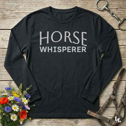 A black long-sleeve t-shirt with 'HORSE WHISPERER' printed in white text across the chest, laid flat on a rustic wooden background with equestrian and floral props.