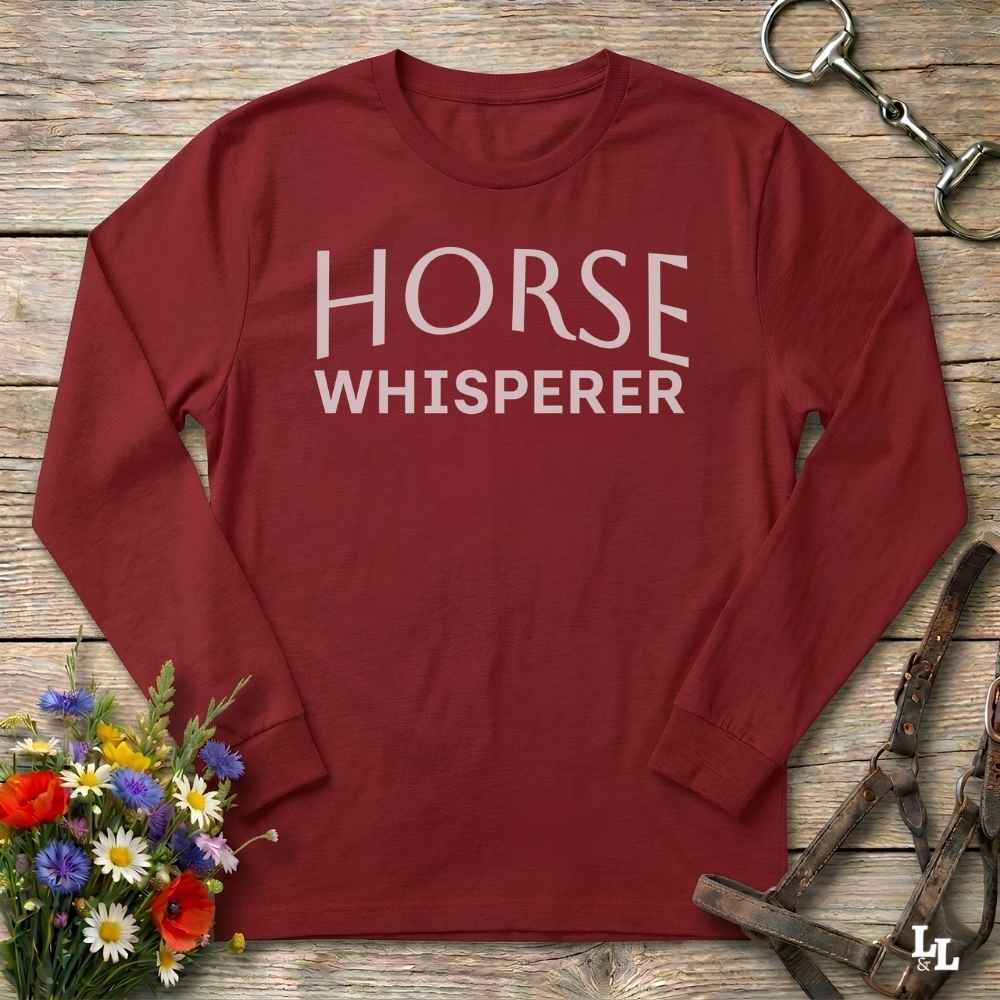 A long-sleeve crewneck shirt in burgundy red with the words 'HORSE WHISPERER' printed in white across the chest, laid flat on a rustic wooden surface with equestrian accessories and flowers around it.