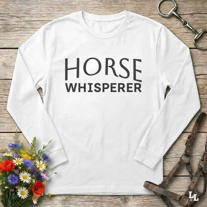 A white long-sleeve t-shirt with 'HORSE WHISPERER' printed in black text across the chest, styled in a flat lay on a rustic wooden background with equestrian accessories and flowers.