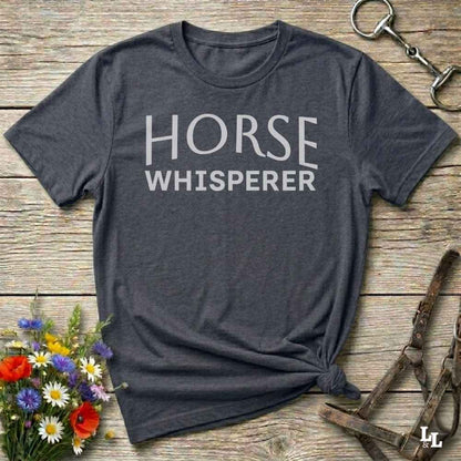 A dark charcoal gray t-shirt with 'HORSE WHISPERER' printed in white text, styled in a flat-lay on a rustic wooden background with equestrian accessories and flowers.