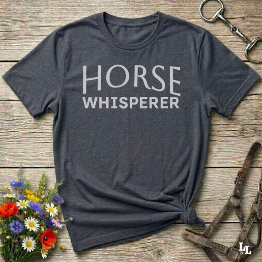 A dark charcoal gray t-shirt with 'HORSE WHISPERER' printed in white text, styled in a flat-lay on a rustic wooden background with equestrian accessories and flowers.