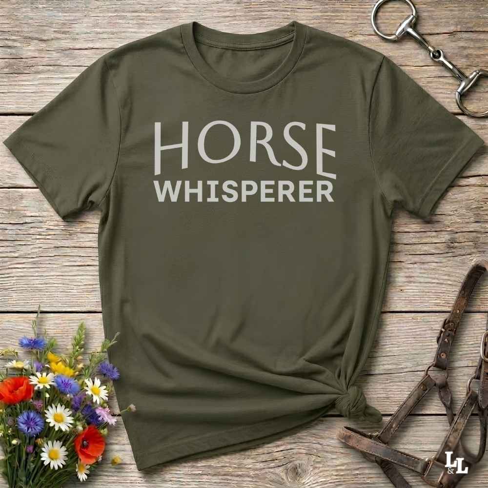 An olive green short-sleeve t-shirt with 'HORSE WHISPERER' printed in white across the chest, styled in a flat-lay on a rustic wooden background with equestrian accessories and flowers.
