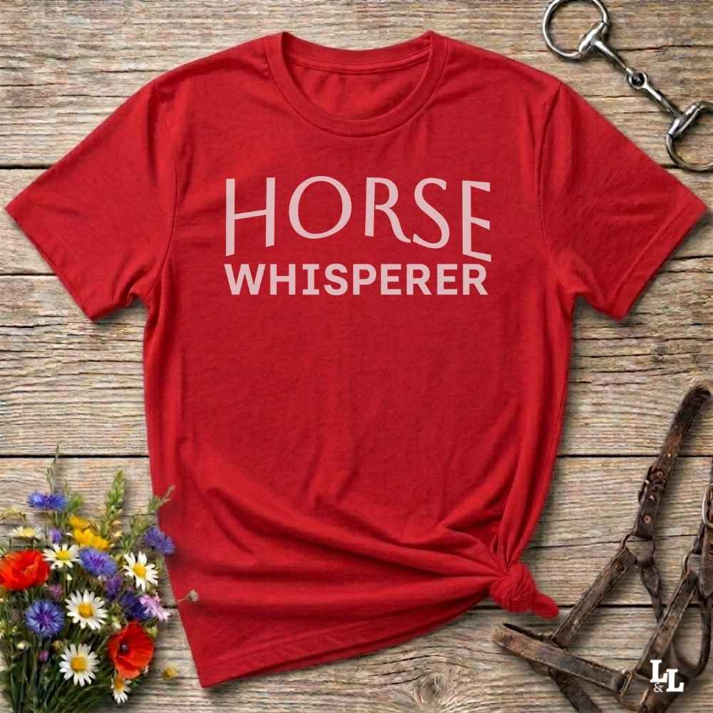 A red short-sleeve t-shirt with 'HORSE WHISPERER' printed in white text across the chest, styled flat on a rustic wooden surface with equestrian accessories and flowers.