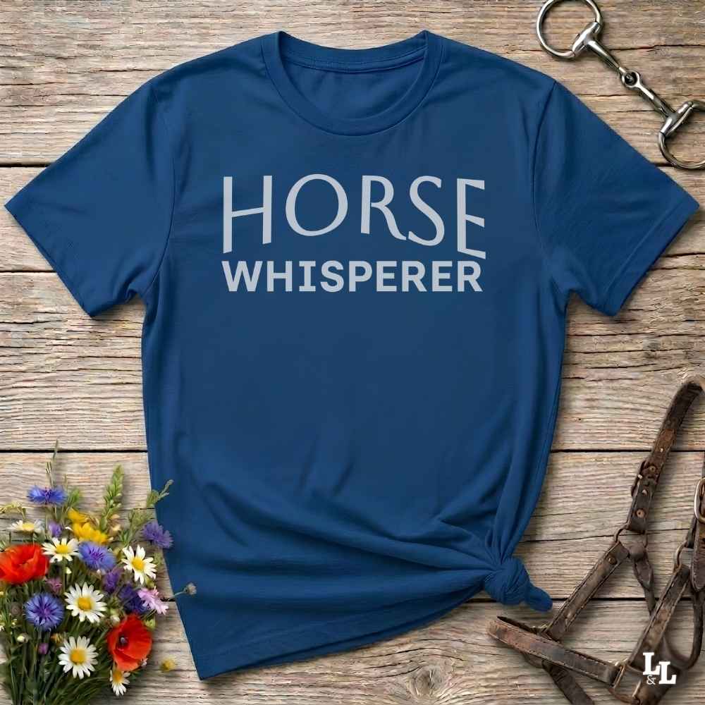 A navy blue short-sleeve t-shirt with 'HORSE WHISPERER' printed in white across the chest, styled flat on a rustic wooden surface with equestrian accessories and flowers.
