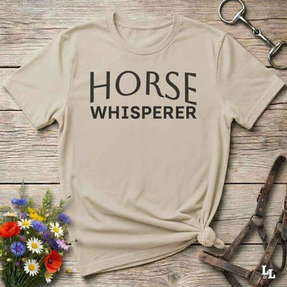 A beige graphic t-shirt with the words 'HORSE WHISPERER' printed in black across the chest, styled in a flat-lay on a rustic wooden background with a horse bit, leather bridle, and a small bouquet of wildflowers.