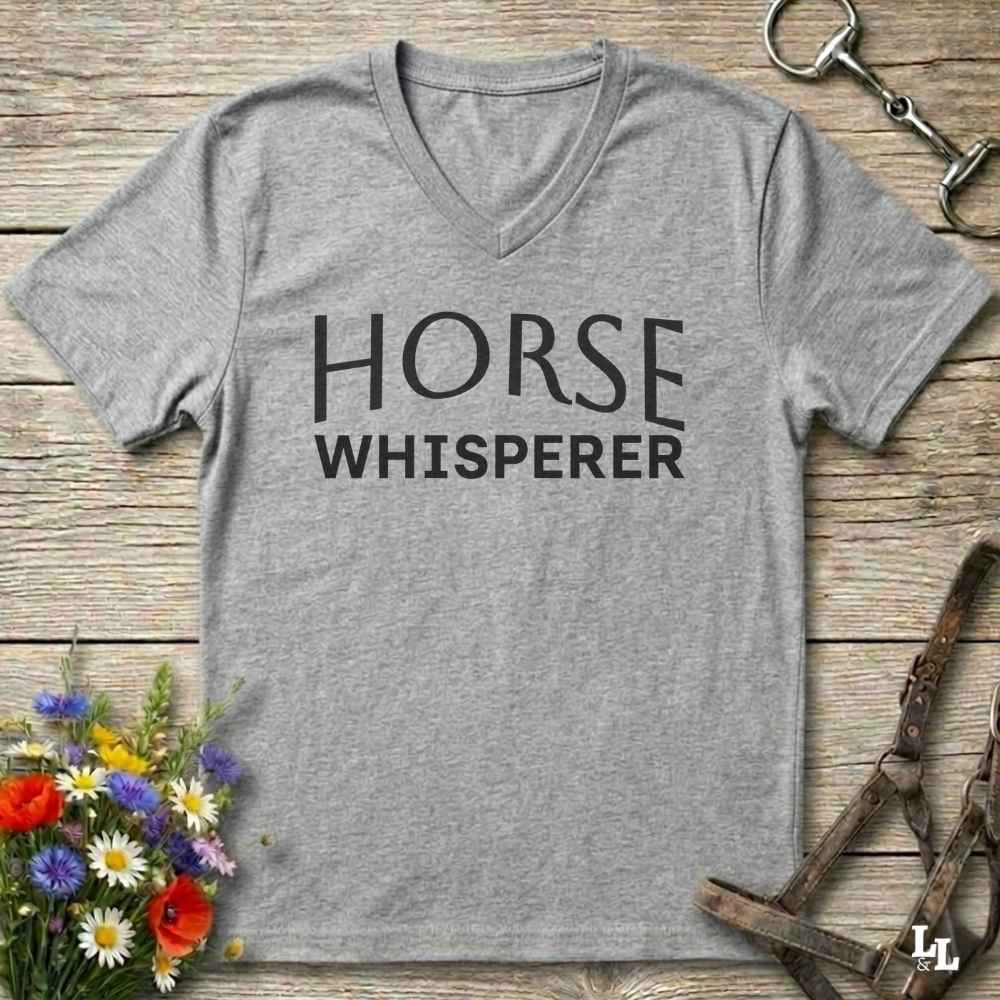 A heather gray V-neck t-shirt with the words 'HORSE WHISPERER' printed in black across the chest, laid flat on a rustic wooden surface with equestrian-themed props including a horse bit, halter, and flowers.