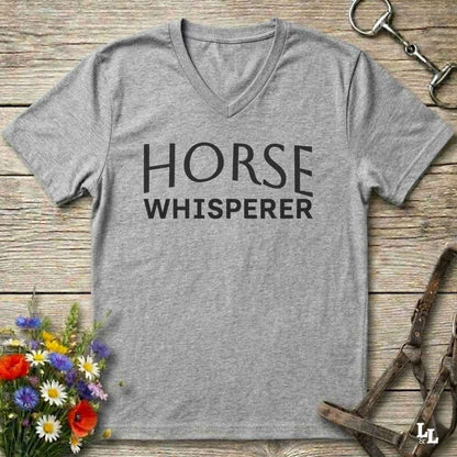 A heather gray V-neck t-shirt with the words 'HORSE WHISPERER' printed in black across the chest, laid flat on a rustic wooden surface with equestrian-themed props including a horse bit, halter, and flowers.