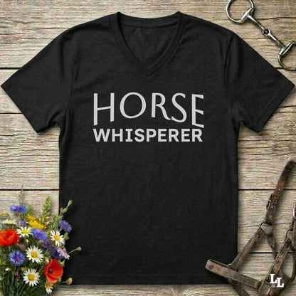 A black V-neck t-shirt with the words 'HORSE WHISPERER' printed in white across the chest, laid flat on a rustic wooden surface with equestrian-themed props including a halter, horse bit, and wildflowers.