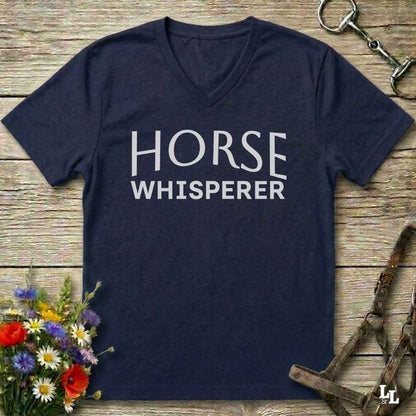 A navy blue V-neck t-shirt with 'HORSE WHISPERER' printed in white text across the chest, laid flat on a rustic wooden surface with equestrian-themed props including a halter, horse bit, and wildflowers.