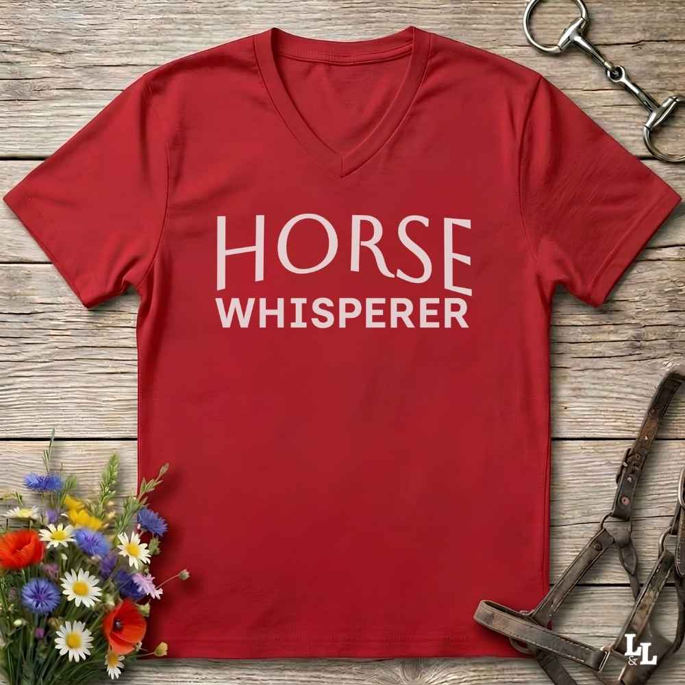 A red V-neck t-shirt with 'HORSE WHISPERER' printed in white text, laid flat on a rustic wooden background with equestrian accessories and flowers.